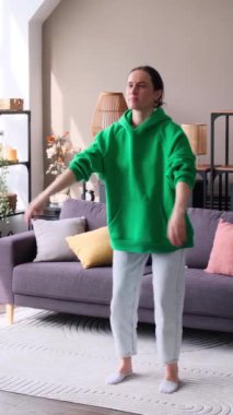 Beyaz, orta yaşlı bir kadın, oturma odasında çömelme egzersizi yapıyor. Ev içi fitness eğitimi ve egzersiz konsepti. Dikey video.