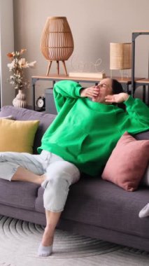 Hoşnutsuz orta yaşlı beyaz kadın esniyor ve esniyor. Oturma odasındaki kanepede dinleniyor. Sıkıntı, hüsran ve acı konsepti. Dikey video.