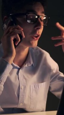 Masada otururken ve akşam ofisinde çalışırken telefona cevap veren, odaklanmış, genç, beyaz, serbest çalışan bir erkek. Dikey video.