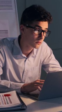 Organize Kafkasyalı genç, yetişkin bir ofis çalışanı dizüstü bilgisayarla evrak işleriyle uğraşıyor, gece yarısı ofisindeki mali evrakları inceliyor. Dikey video.