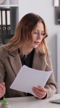Konsantre olmuş, beyaz, genç, yetişkin bir ofis çalışanı çevrimiçi video görüşmesi yapıyor ve ofiste belgelerle çalışıyor. Dikey video.