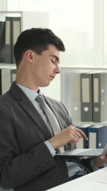 Modern ofiste dizüstü bilgisayarla çalışan, düşünen ve geliştiren düşünceli, genç, beyaz bir erkek işçi. Dikey video.