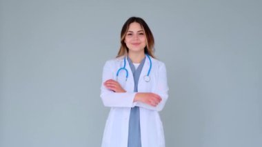 Gülümseyen ve rahat bir beyaz kadın doktor ya da stajyer kadının portresi gri arka planda çapraz kollarla duruyor. Mutlu profesyonel hemşire.