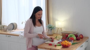 Orta yaşlı, beyaz, hamile bir kadın ev mutfağında yemek hazırlıyor ve bıçakla sağlıklı salata için domates kesiyor..