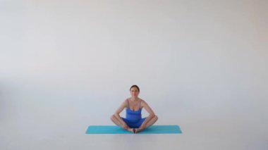 Sportif ve hamile orta yaşlı beyaz kadın yoga seansından zevk alıyor, Lotus pozisyonunda beyaz arka planda oturuyor..