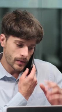 Odaklanmış orta yaşlı Kafkasyalı işadamı telefonla konuşuyor ve ofiste belgelerle çalışıyor. Evrak işleri ve sohbet anlayışı. Dikey video.