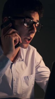 Stern Kafkasyalı genç yetişkin ofis çalışanı akşam ofisinde çalışırken müşteri ya da iş arkadaşlarıyla telefon görüşmelerinde bulundu. Dikey video.