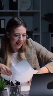 Beyaz, genç, pozitif, yetişkin bir kadın, evrak işi yapıyor ve akşam ofisinde çalışırken müzik eşliğinde dans ediyor. Dikey video.