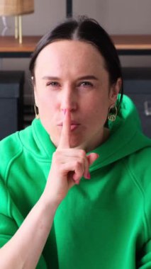 Ciddi, beyaz, orta yaşlı bir kadın sessiz bir jest yapıyor, parmakları dudaklarında, evdeki kanepede oturuyor. Mahremiyet ve gizlilik sembolü. Dikey video.