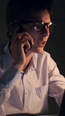 Ciddi bir Kafkas erkek çalışanı gece geç saatlerde çalışırken müşterisiyle konuşmak için akıllı telefonu kullanıyor. Dikey video.