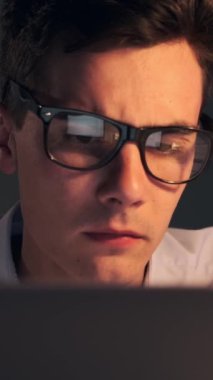 Akşam ofisinde dizüstü bilgisayarla çalışan Kafkasyalı genç bir erkek ofis çalışanının portresini yakından çek. Dikey video.