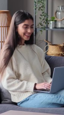 Neşeli genç beyaz kadın internette sosyal medyada sohbet ediyor, dizüstü bilgisayar kullanıyor ve gülüyor, hafta sonları evde boş vakit geçiriyor. Dikey video.