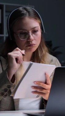 Konsantre olmuş Kafkasyalı genç, yetişkin kadın ofis çalışanı. Not alıyor ve akşam ofisindeki internet üzerinden konuşuyor. Dikey video.