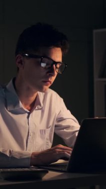 Yorgun, genç, beyaz bir erkek, dizüstü bilgisayarla çalışıyor ve gece yarısı ofisinde hesap özetleri hazırlıyor. Dikey video.