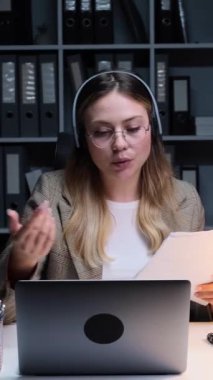 Ofisteki çevrimiçi video görüşmesi sırasında dizüstü bilgisayar ve belgelerle çalışan Kafkasyalı genç bir iş kadını. Dikey video.