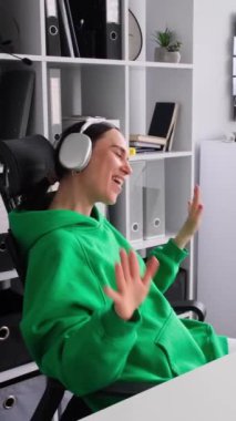 Güler yüzlü beyaz kadın kulaklıkla müzik dinliyor, ofiste telefon kullanıyor, teneffüste boş vakit geçiriyor. Dikey video.