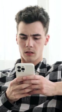 Akıllı telefon kullanan ve evde internet gezerken facepalm gösteren Kafkasyalı genç adam memnun değil ve üzgün. Dikey video.