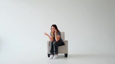 Olumlu, beyaz, genç, yetişkin bir kadın akıllı telefon kullanarak konuşma yapıyor ve beyaz arka planda koltukta dinleniyor. Uzak bağlantı ve iletişim.