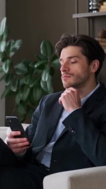 Ciddi ve düşünceli Kafkas erkek işadamı cep telefonunu internette gezinmek için kullanıyor, koltukta oturuyor ve evde çalışıyor. Dikey video.