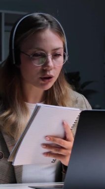 Konsantre olmuş Kafkasyalı genç yetişkin kadın çalışan akşam ofisinde çalışırken video görüşmeleri sırasında not alıyor. Dikey video.