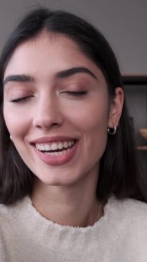 Gülümseyen genç, beyaz bir kadın çevrimiçi video görüşmesi yapıyor, telefonu tutuyor, web kamerasıyla konuşuyor ve evdeki kameraya bakıyor. Dikey video.