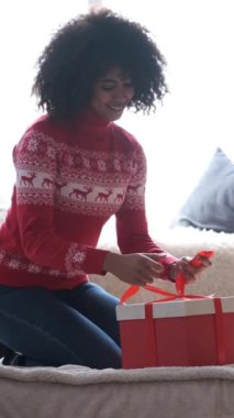 Afrika kökenli Amerikalı kadın evde Noel hediyesi kutusuna saten kurdele bağlıyor.