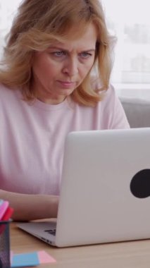 Neşeli, orta yaşlı, beyaz, uzaktan kumandalı bir kadın. Laptop kullanıyor ve ev ofisinden çalışıyor. İş planlama ve geliştirme. Dikey video.