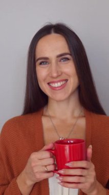 Beyaz arka planda duran, çay içen, gülümseyen ve kameraya bakan orta yaşlı beyaz bir kadının portresi. Dikey video.