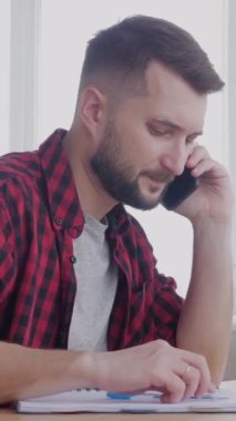 Merkez büroda çalışan, notlar alan ve telefon görüşmeleri sırasında iş görüşmelerine katılan orta yaşlı, odaklı beyaz bir adam. Dikey video.