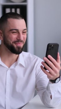 Ofiste çalışan orta yaşlı beyaz bir adam var ve cep telefonu kullanarak internet üzerinden görüşme ya da danışmanlık yapıyor. Dikey video.