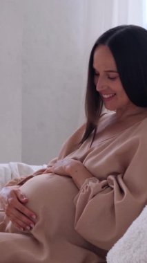 Pozitif orta yaşlı, beyaz hamile bir kadın evdeki kanepede bebek bekliyor. Koruma ve rahatlama kavramı. Dikey video.