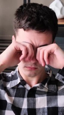 Kafkas asıllı genç bir adamın portresi. Gözlerini ovuşturuyor, evdeki kanepede oturuyor. Yorgunluk, yorgunluk ve yorgunluk konsepti. Dikey video.