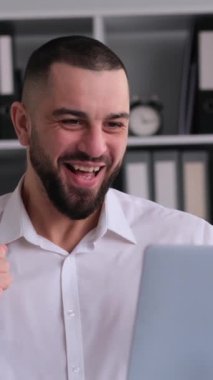 Ofiste dizüstü bilgisayarla çalışırken zafer ya da iyi haberi kutlayan başarılı beyaz bir adamın portresi. Dikey video.