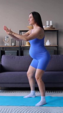 Neşeli, orta yaşlı, hamile, beyaz bir kadın hamilelik ya da evde egzersiz yapmak için çömeliyor. Sağlıklı bir anne olma kavramı. Dikey video.