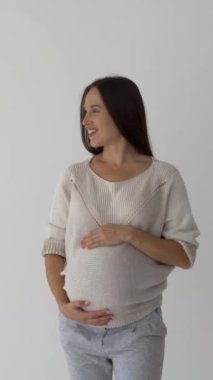 Neşeli hamile bir kadının portresi beyaz arka planda bebek çarpması, mutluluk, sıcaklık ve annelik şefkati yayıyor. Dikey video.