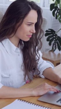 Kafkasyalı mutlu orta yaşlı kadın internette çalışırken ya da çalışırken dizüstü bilgisayar kullanıyor ve evdeki oturma odasında ya da ofiste not defterine yazıyor. Dikey video.