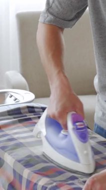 Evde sabahları demir kullanan orta yaşlı, mutlu bir beyaz adam. Kumaş ütüleme ve temizlik konsepti. Dikey video.