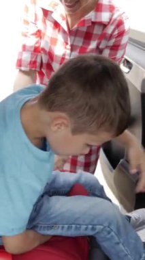 Anne, oğlunu dikkatli bir şekilde bir aracın içindeki çocuk koltuğuna bağlıyor ve yolculuktan önce güvenliğini sağlıyor. Dikey video.
