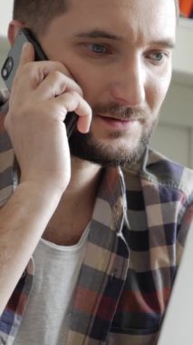 Beyaz, orta yaşlı bir erkeğin portresi. Uzaktan kumandalı ciddi bir işçi. Evde dizüstü bilgisayarla çalışırken cep telefonu kullanıyor. Dikey video.