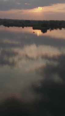 Gün batımının sıcak renkleriyle aydınlanan bir nehir suyunun havadan görünüşü, sakin bir akşam manzarasında etrafındaki doğayla güzel bir şekilde harmanlanıyor. Dikey video.