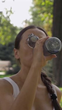 Şehir parkında egzersiz yaptıktan sonra susuzluk, sağIık ve doğum öncesi tedaviye odaklanarak su içen arkadaş canlısı hamile bir kadın. Dikey video.