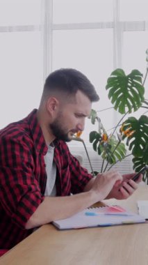Ciddi bir orta yaşlı beyaz erkek çalışanı cep telefonuyla çalışıyor ve evdeki oturma odasında not defterine not alıyor. Dikey video.