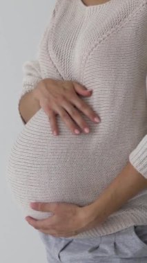 Hamile bir kadının, beyaz bir arka planda bebek şişliğini okşarken, bakım, şefkat, hamilelik ve annelik beklentilerini ifade ederken. Dikey video.