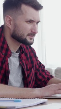 Konsantre olmuş Kafkasyalı orta yaşlı adam online olarak çalışıyor ya da öğreniyor dizüstü bilgisayarla, evde oturma odasında masa başında oturuyor. Dikey video.