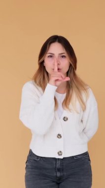 Resmi Kafkasyalı genç bir kadın. Bej arka planda dudaklarında parmak olan sessiz bir jest yapıyor. Sır kavramı. Dikey video.