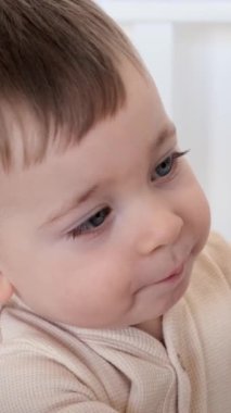 Evdeki oyun odasında beşikte oturan, tahta oyuncakla oynayan mutlu ve gülümseyen küçük beyaz bir çocuğun portresi. Dikey video.