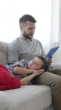 Kafkasyalı küçük bir çocuk ve orta yaşlı bir baba birlikte kitap okuyor, aile eğlencesini evde oturma odasındaki kanepede dinlenerek geçiriyor. Dikey video.