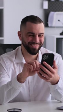 Heyecanlı beyaz, orta yaşlı erkek çalışanın tatilde cep telefonunu ofiste boş vakit geçirmek ve eğlenmek için kullanıyor. Dikey video.