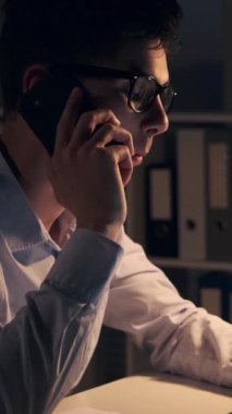 Konsantre olmuş Kafkasyalı genç, yetişkin iş adamı akşam ofisinde çalışırken telefonla konuşuyor. Sohbet ya da pazarlık kavramı. Dikey video.