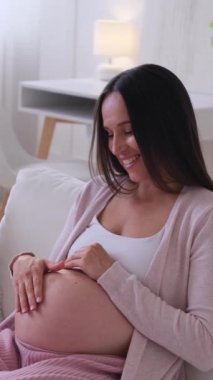 Heyecanlı beyaz orta yaşlı kadın hamile karnına dokunuyor, evdeki kanepede dinleniyor. Hamilelik, sağlık ve koruma kavramı. Dikey video.